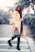 A fashionable woman wearing a long camel-colored coat, black knee-high boots, and holding a stylish handbag is standing on outdoor steps. The background includes trees and a building, creating an urban environment.