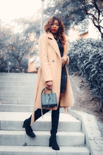 A fashionable woman wearing a long camel-colored coat, black knee-high boots, and holding a stylish handbag is standing on outdoor steps. The background includes trees and a building, creating an urban environment.