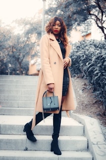 A fashionable woman wearing a long camel-colored coat, black knee-high boots, and holding a stylish handbag is standing on outdoor steps. The background includes trees and a building, creating an urban environment.