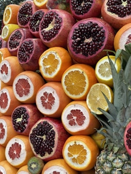 A vibrant assortment of halved fruits including pomegranates, oranges, grapefruits, lemons, and a pineapple. The arrangement highlights the fruits' colorful insides, with pomegranate seeds glistening. The scene is lush and fresh, emphasizing the natural beauty and variety of the fruits.