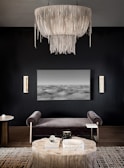 An elegant, modern living room with dark walls features a large, intricate chandelier with cascading elements as its focal point. Below, a gray velvet bench sits in front of a black-and-white abstract landscape artwork. A round wooden table is placed on a patterned rug, adorned with decorative objects, including a book and sculptural items. On the side, two vertical wall sconces add a touch of warmth and light to the space.