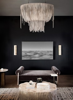 An elegant, modern living room with dark walls features a large, intricate chandelier with cascading elements as its focal point. Below, a gray velvet bench sits in front of a black-and-white abstract landscape artwork. A round wooden table is placed on a patterned rug, adorned with decorative objects, including a book and sculptural items. On the side, two vertical wall sconces add a touch of warmth and light to the space.