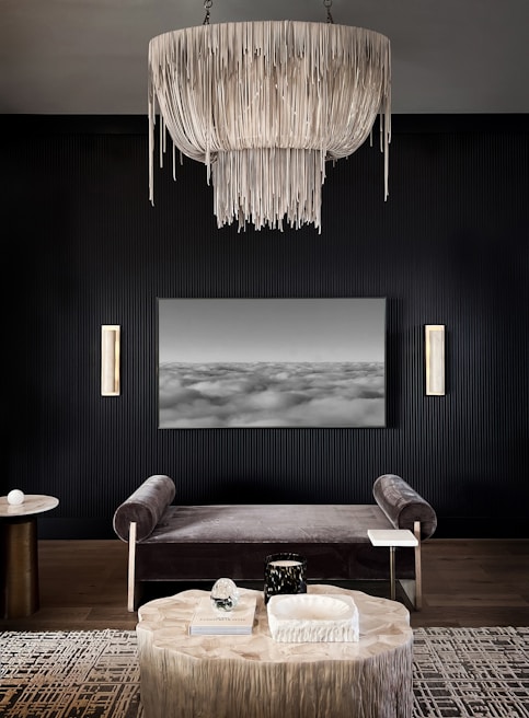 An elegant, modern living room with dark walls features a large, intricate chandelier with cascading elements as its focal point. Below, a gray velvet bench sits in front of a black-and-white abstract landscape artwork. A round wooden table is placed on a patterned rug, adorned with decorative objects, including a book and sculptural items. On the side, two vertical wall sconces add a touch of warmth and light to the space.