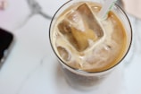A glass filled with iced coffee containing large ice cubes. A straw with a green design is inserted into the drink. The surface is creamy, suggesting that milk or cream has been added. The background features a marble texture, and a partial view of a smartphone is visible at the edge.