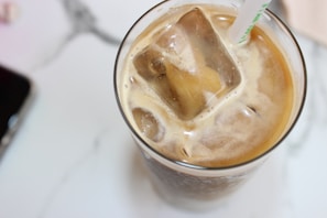A glass filled with iced coffee containing large ice cubes. A straw with a green design is inserted into the drink. The surface is creamy, suggesting that milk or cream has been added. The background features a marble texture, and a partial view of a smartphone is visible at the edge.