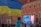 A crowd gathered outside in front of a large screen displaying a person's image and text advertising remarks by a president in Warsaw, Poland. The setting is against a large building with a red facade. A waving yellow and blue flag is prominent in the foreground.