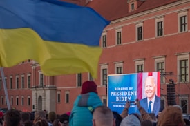 A crowd gathered outside in front of a large screen displaying a person's image and text advertising remarks by a president in Warsaw, Poland. The setting is against a large building with a red facade. A waving yellow and blue flag is prominent in the foreground.