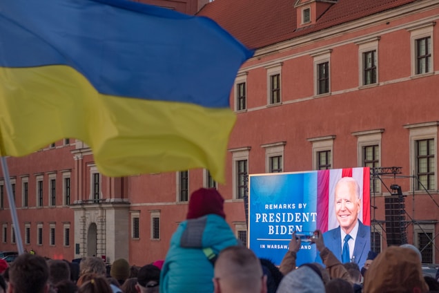 A crowd gathered outside in front of a large screen displaying a person's image and text advertising remarks by a president in Warsaw, Poland. The setting is against a large building with a red facade. A waving yellow and blue flag is prominent in the foreground.