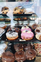 An assortment of pastries displayed in a glass case, including various types of donuts and pastries with different toppings like chocolate, pink frosting, and sprinkles. Some pastries are dusted with powdered sugar, while others feature chocolate chips.