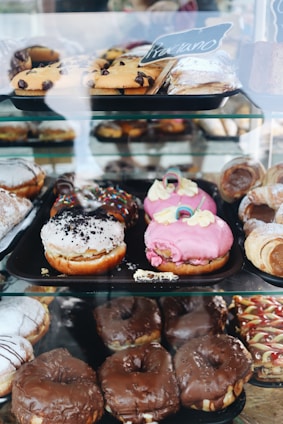 An assortment of pastries displayed in a glass case, including various types of donuts and pastries with different toppings like chocolate, pink frosting, and sprinkles. Some pastries are dusted with powdered sugar, while others feature chocolate chips.