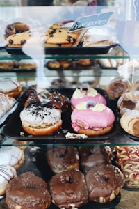 An assortment of pastries displayed in a glass case, including various types of donuts and pastries with different toppings like chocolate, pink frosting, and sprinkles. Some pastries are dusted with powdered sugar, while others feature chocolate chips.