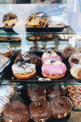 An assortment of pastries displayed in a glass case, including various types of donuts and pastries with different toppings like chocolate, pink frosting, and sprinkles. Some pastries are dusted with powdered sugar, while others feature chocolate chips.