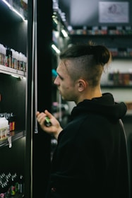 A person with a shaved hairstyle is standing next to shelves filled with small bottles, possibly in a store that sells e-liquids or vaping products. The individual is holding one of the small bottles while looking at the shelves with a focused expression. The setting is dimly lit, highlighting the rows of colorful bottles under soft lighting.