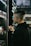 A person with a shaved hairstyle is standing next to shelves filled with small bottles, possibly in a store that sells e-liquids or vaping products. The individual is holding one of the small bottles while looking at the shelves with a focused expression. The setting is dimly lit, highlighting the rows of colorful bottles under soft lighting.