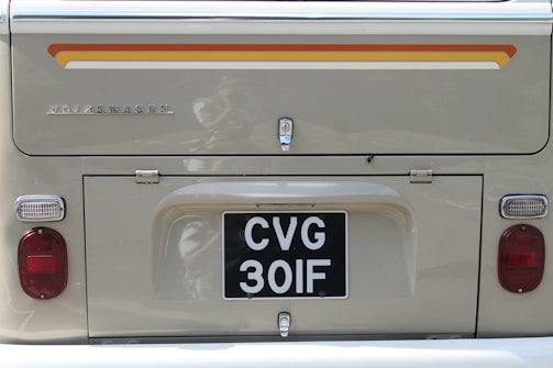 The rear side of a classic Volkswagen vehicle is visible, featuring a retro design with a beige color. A decorative stripe in orange and yellow runs horizontally near the top. The license plate reads 'CVG 30IF'. The vehicle shows metal and chrome detailing, with two red circular lights on either side.