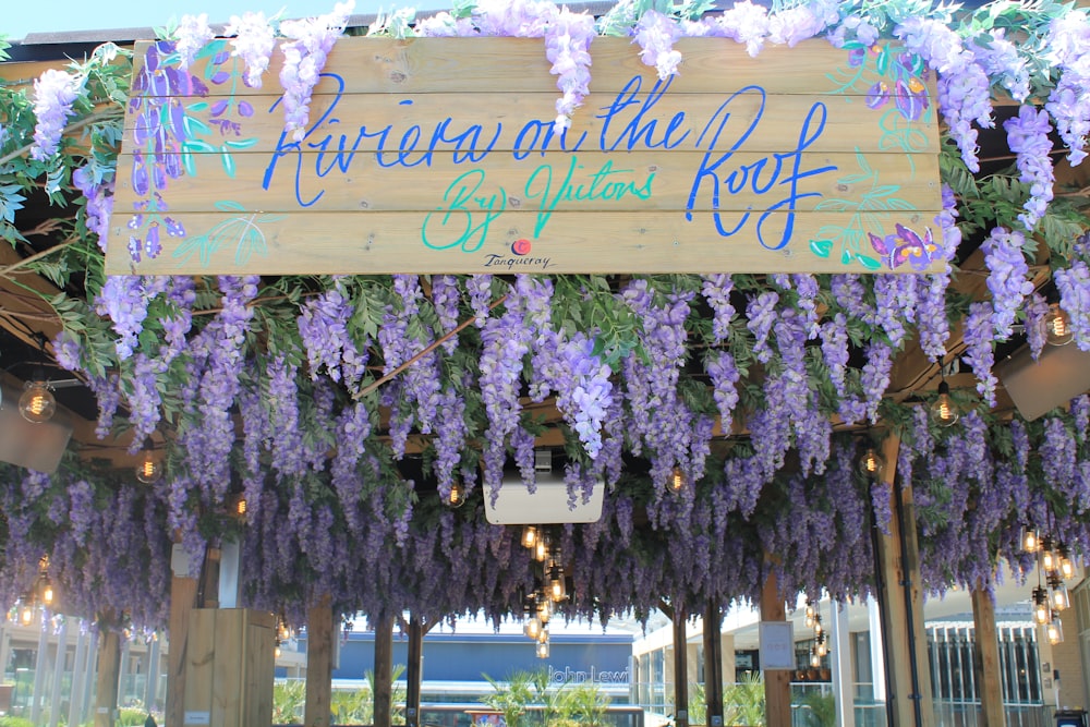 a wooden sign hanging from the side of a building covered in purple flowers