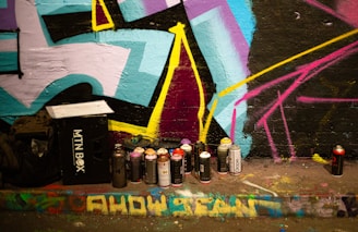 A vibrant street art scene with a wall covered in colorful graffiti. Spray paint cans of various colors are scattered on the ground, next to a black MTN box. The wall features bold geometric shapes in shades of blue, pink, yellow, and black. A few bags are placed next to the MTN box.