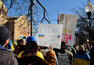 A group of people gathered outdoors holding signs with messages of support for Ukraine. The setting includes a church building in the background under a clear blue sky. The scene is vibrant with Ukrainian flags and a prominent sign that reads 'Chicago stands with Ukraine!'