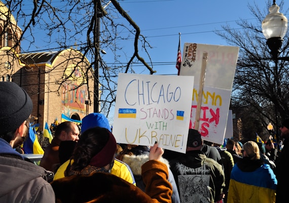 A group of people gathered outdoors holding signs with messages of support for Ukraine. The setting includes a church building in the background under a clear blue sky. The scene is vibrant with Ukrainian flags and a prominent sign that reads 'Chicago stands with Ukraine!'