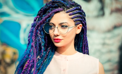 A person with braided hair in vibrant shades of blue and purple, wearing large round glasses and a light pink sleeveless top. The background is a colorful urban mural.