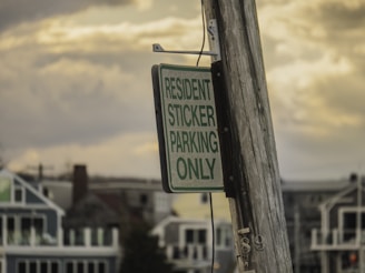 A wooden pole supports a sign that reads 'Resident Sticker Parking Only' in front of a row of houses. The sky is overcast, with a mix of cloud coverage in the background.