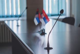 A conference table with microphones and two small flags on stands. The room appears to be well-lit with natural light coming from a window. The flags have a red, blue, and white design. The focus is on the microphones which are placed on the glossy surface of the table.