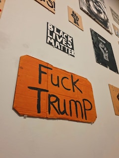 A collection of protest signs on a wall, including one prominently featuring an orange cardboard with bold black text expressing opposition to Trump. Other signs display messages like 'Black Lives Matter' and feature symbolic imagery such as a raised fist.