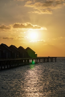A serene sunset scene with the sun low in the sky, casting a golden glow over the water. Overwater bungalows are positioned along a pier, silhouetted against the vibrant sky. The sea reflects the warm tones of the setting sun, adding to the tranquil atmosphere.