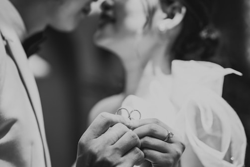 A close-up black and white photograph of a couple, focusing on their hands holding wedding rings. The faces are slightly blurred in the background, emphasizing the intimacy of the moment. The man is in a suit and the woman is in a bridal gown with delicate fabric details.