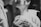 A close-up black and white photograph of a couple, focusing on their hands holding wedding rings. The faces are slightly blurred in the background, emphasizing the intimacy of the moment. The man is in a suit and the woman is in a bridal gown with delicate fabric details.