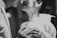 A close-up black and white photograph of a couple, focusing on their hands holding wedding rings. The faces are slightly blurred in the background, emphasizing the intimacy of the moment. The man is in a suit and the woman is in a bridal gown with delicate fabric details.
