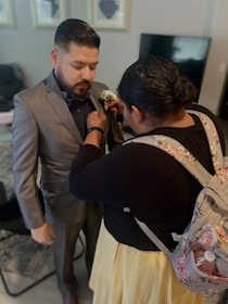 A man in a formal suit is having a boutonniere pinned to his jacket by a woman. The setting is a living room with a television in the background and framed artwork on the walls. The woman is wearing a floral backpack and a yellow skirt.
