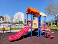 A vibrant playground set with several slides and climbing structures in red, yellow, and blue is surrounded by a cityscape of tall buildings. The area is bright and sunny, with a clear blue sky and a few trees.