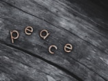 Wooden letters spelling out 'peace' are positioned on a textured, weathered wooden surface. The letters stand out with a light, warm color against the darker, grayish tones of the wood.