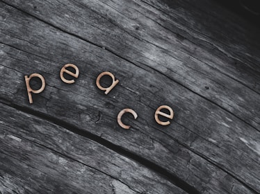 Wooden letters spelling out 'peace' are positioned on a textured, weathered wooden surface. The letters stand out with a light, warm color against the darker, grayish tones of the wood.