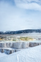A stunning landscape features terraces of geothermal formations covered in snow, with vibrant colors of yellow and green contrasting against the white layers. The background displays a range of snow-covered mountains under a cloudy sky, and steam can be seen rising from parts of the landscape.