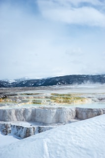 A stunning landscape features terraces of geothermal formations covered in snow, with vibrant colors of yellow and green contrasting against the white layers. The background displays a range of snow-covered mountains under a cloudy sky, and steam can be seen rising from parts of the landscape.