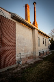 A building with brick and stone exterior features a prominent chimney and an exhaust pipe on its roof. The structure has aged, evident in its weathered appearance and faded paint. The surroundings include a patch of grass and some shrubs lining the side of the building.