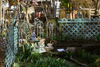 A garden setting with a variety of elements including trellises, a bird bath with a decorative bird on top, and a miniature house. There are flowering plants and greenery around, with lattice fences and garden ornaments such as a metallic sphere and a woven hanging decoration. The scene is bathed in sunlight, highlighting the vibrant colors and textures.