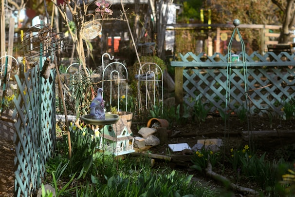 A garden setting with a variety of elements including trellises, a bird bath with a decorative bird on top, and a miniature house. There are flowering plants and greenery around, with lattice fences and garden ornaments such as a metallic sphere and a woven hanging decoration. The scene is bathed in sunlight, highlighting the vibrant colors and textures.
