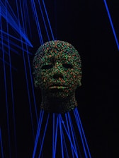 A head sculpture is covered in vibrant multicolored dots, illuminated by a blacklight. Surrounding the head are glowing blue lines, creating a surreal and futuristic atmosphere.