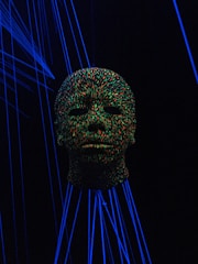 A head sculpture is covered in vibrant multicolored dots, illuminated by a blacklight. Surrounding the head are glowing blue lines, creating a surreal and futuristic atmosphere.