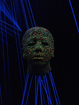 A head sculpture is covered in vibrant multicolored dots, illuminated by a blacklight. Surrounding the head are glowing blue lines, creating a surreal and futuristic atmosphere.