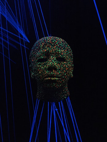 A head sculpture is covered in vibrant multicolored dots, illuminated by a blacklight. Surrounding the head are glowing blue lines, creating a surreal and futuristic atmosphere.