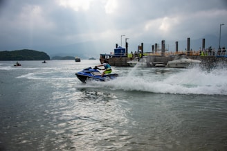 A person is riding a jet ski on the water, creating a trail of white splash behind them. There is a dock in the background with several boats moored and a small crowd of people. The scene is set under a cloudy sky with distant mountains partially visible.