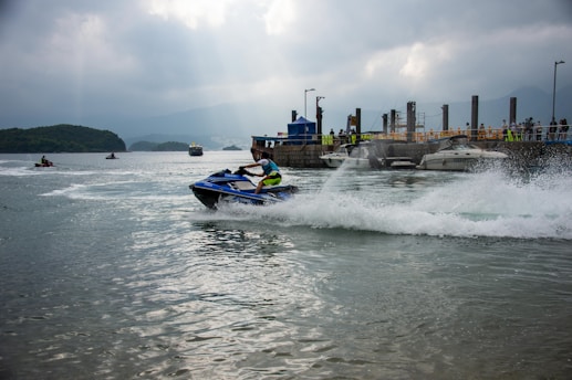 A person is riding a jet ski on the water, creating a trail of white splash behind them. There is a dock in the background with several boats moored and a small crowd of people. The scene is set under a cloudy sky with distant mountains partially visible.