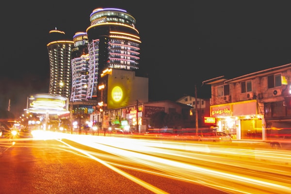 A nighttime city scene with modern, illuminated skyscrapers featuring distinct architectural designs. Bright light trails from passing vehicles on a busy street add dynamic motion to the image. Surrounding buildings display vibrant neon lights and signage, contributing to the lively urban atmosphere.