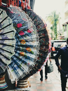 An array of ornate, hand-painted fans with floral patterns are on display, hanging by red clips against an outdoor city background. The fans exhibit a variety of colors and intricate details, including lace edges. A street scene is visible in the blurry background, with a person walking and cars passing by.