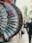 An array of ornate, hand-painted fans with floral patterns are on display, hanging by red clips against an outdoor city background. The fans exhibit a variety of colors and intricate details, including lace edges. A street scene is visible in the blurry background, with a person walking and cars passing by.