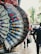 An array of ornate, hand-painted fans with floral patterns are on display, hanging by red clips against an outdoor city background. The fans exhibit a variety of colors and intricate details, including lace edges. A street scene is visible in the blurry background, with a person walking and cars passing by.